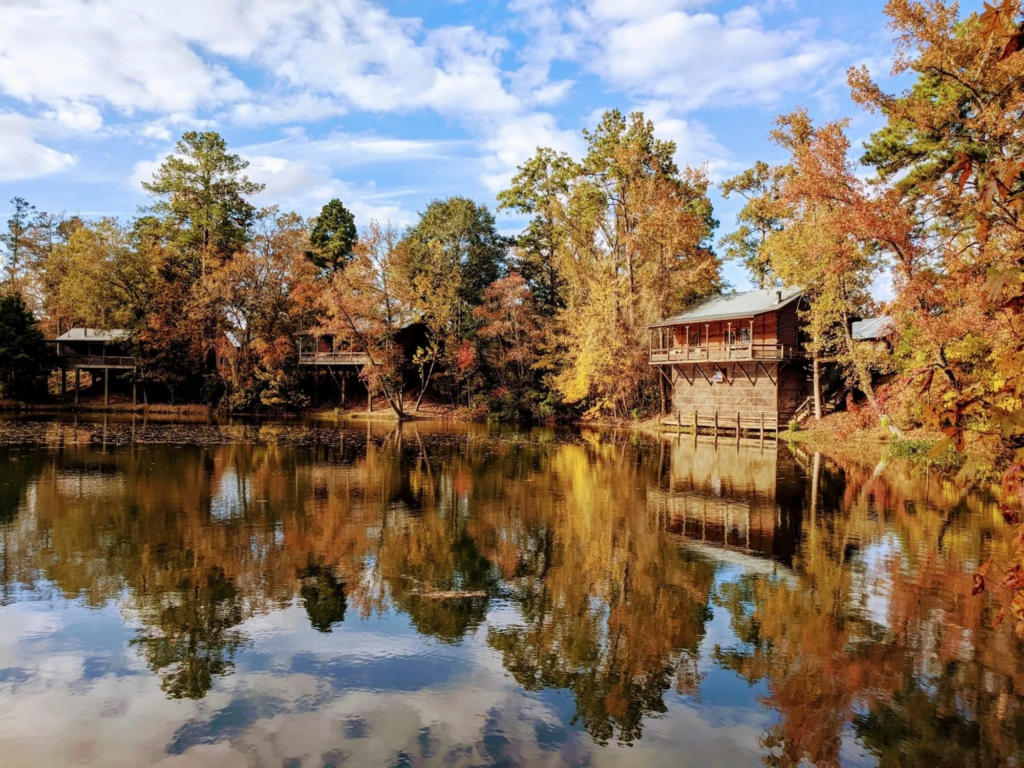Log cabins and trees reflected in a lake at the Retreat at Artesian Lakes