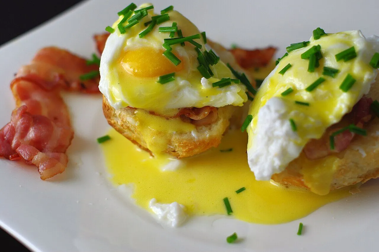 Two eggs Benedict topped with hollandaise and chopped chives, with bacon on the plate
