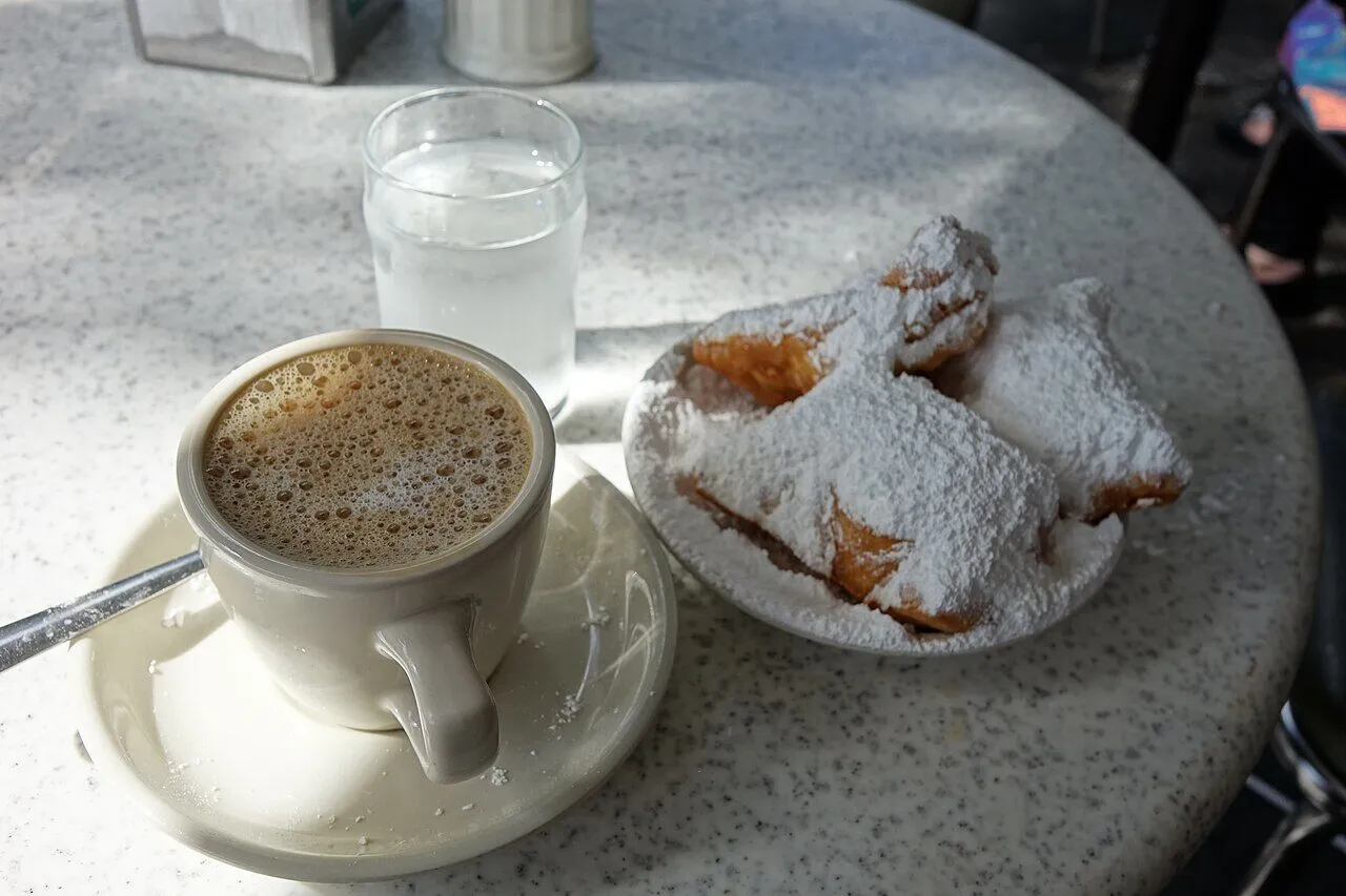 A cup of cafe au lait beside powdered-sugar beignets on a cafe table