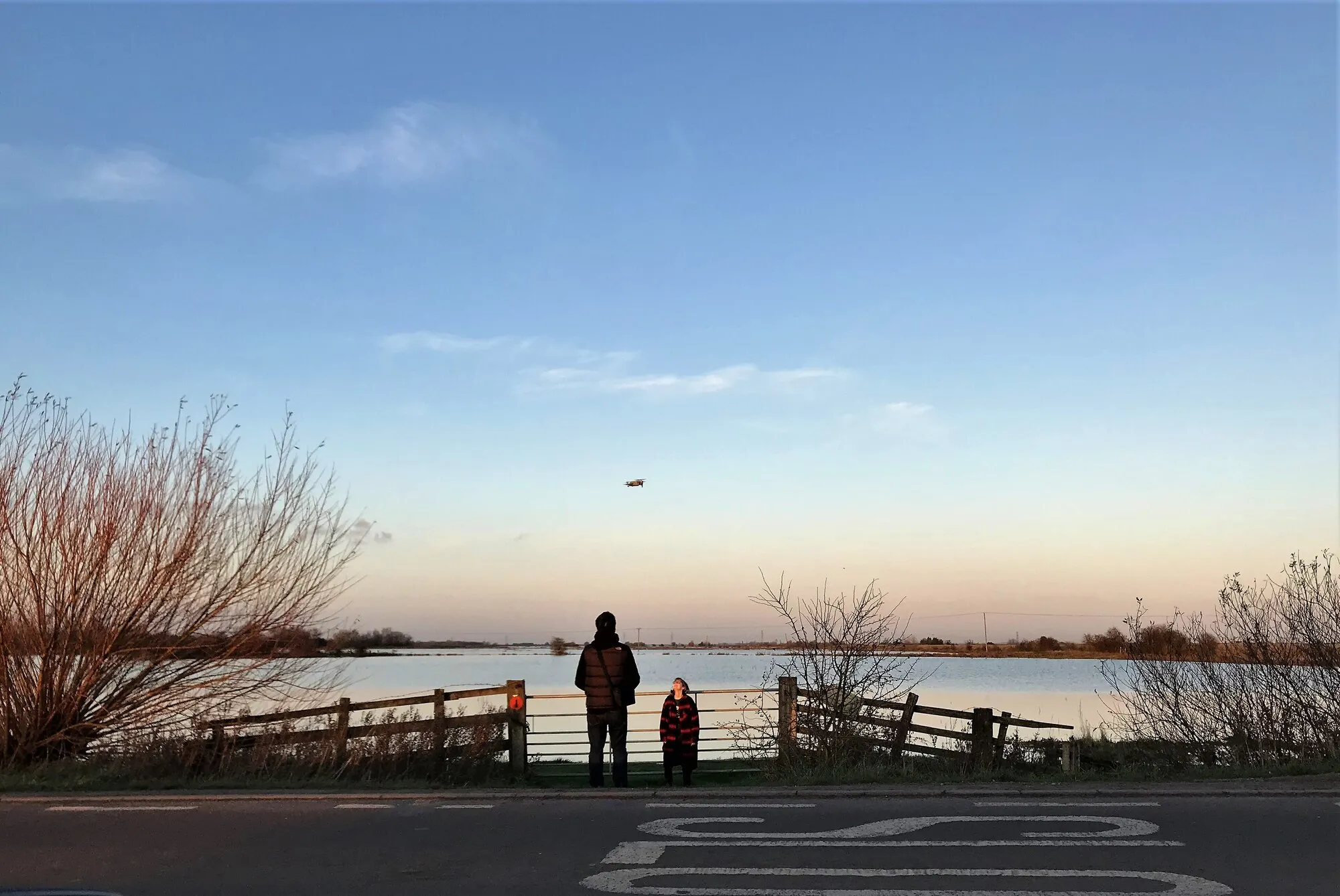 A survey pilot holding a drone controller beside flooded fields while a small quadcopter flies overhead