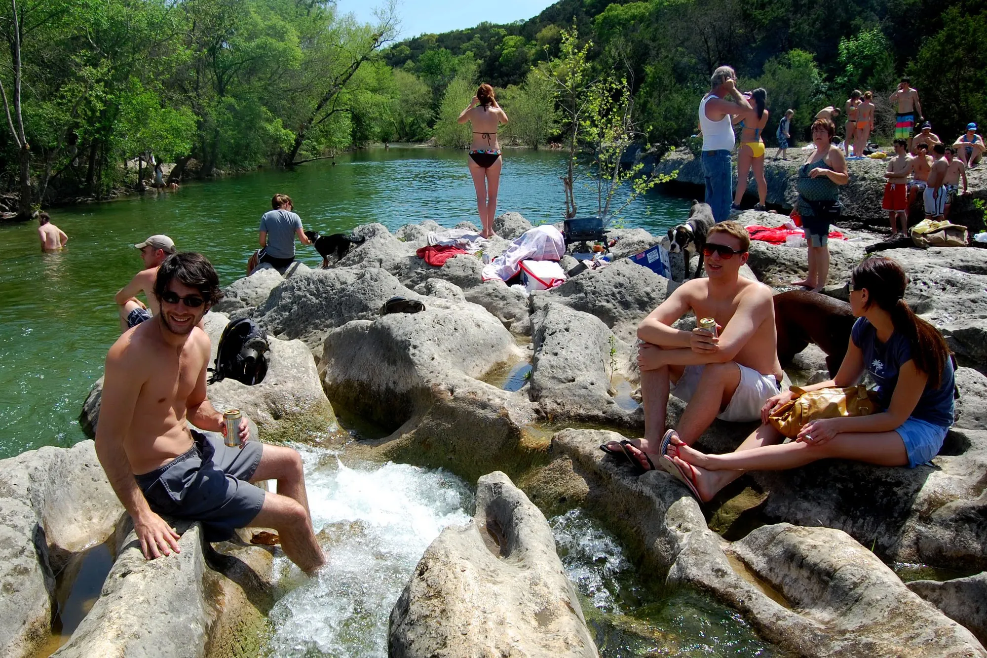 Swimmers resting on limestone ledges beside the green water at Sculpture Falls on Barton Creek Greenbelt