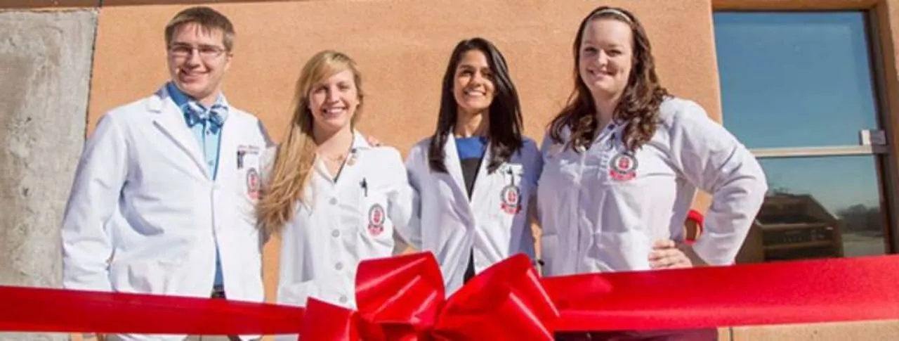 Four Paul L. Foster medical students in white coats standing behind a red ribbon outside a clinic