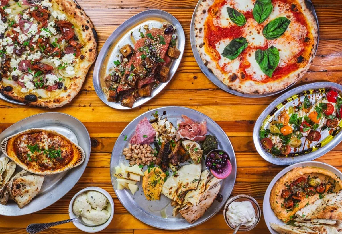 An overhead spread of Il Forno pizzas, roasted vegetables, bread and small plates on a wooden table