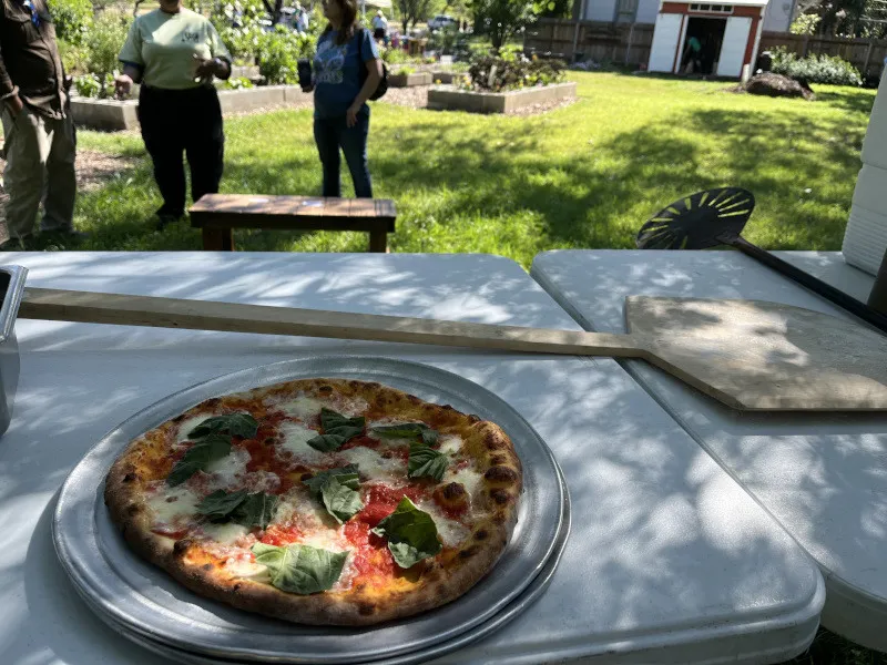 A Margherita pizza on a metal tray at an outdoor Il Forno event, with garden beds behind it