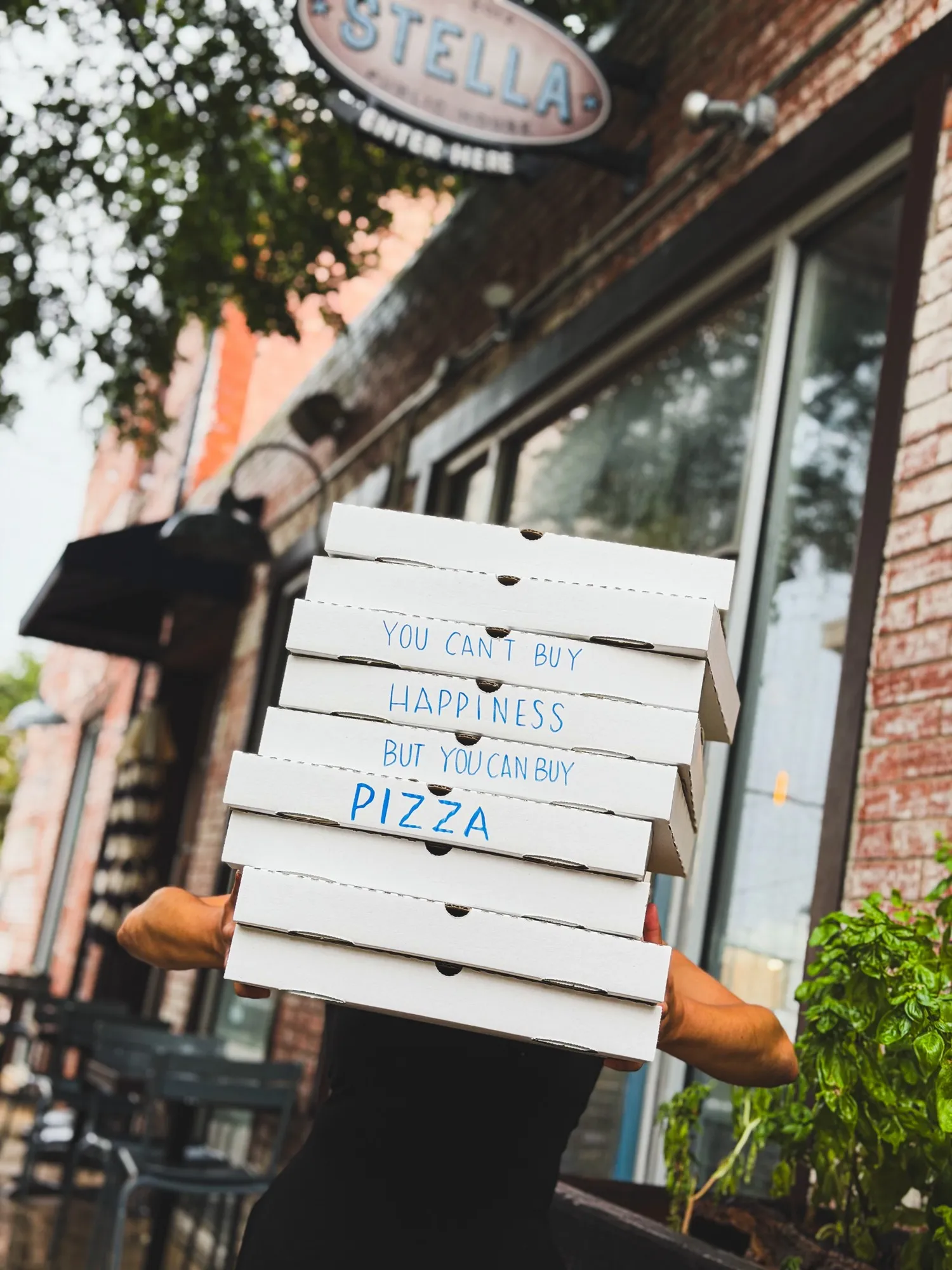 A Stella Public House staff member carrying a tall stack of pizza boxes beneath the restaurant's oval sign