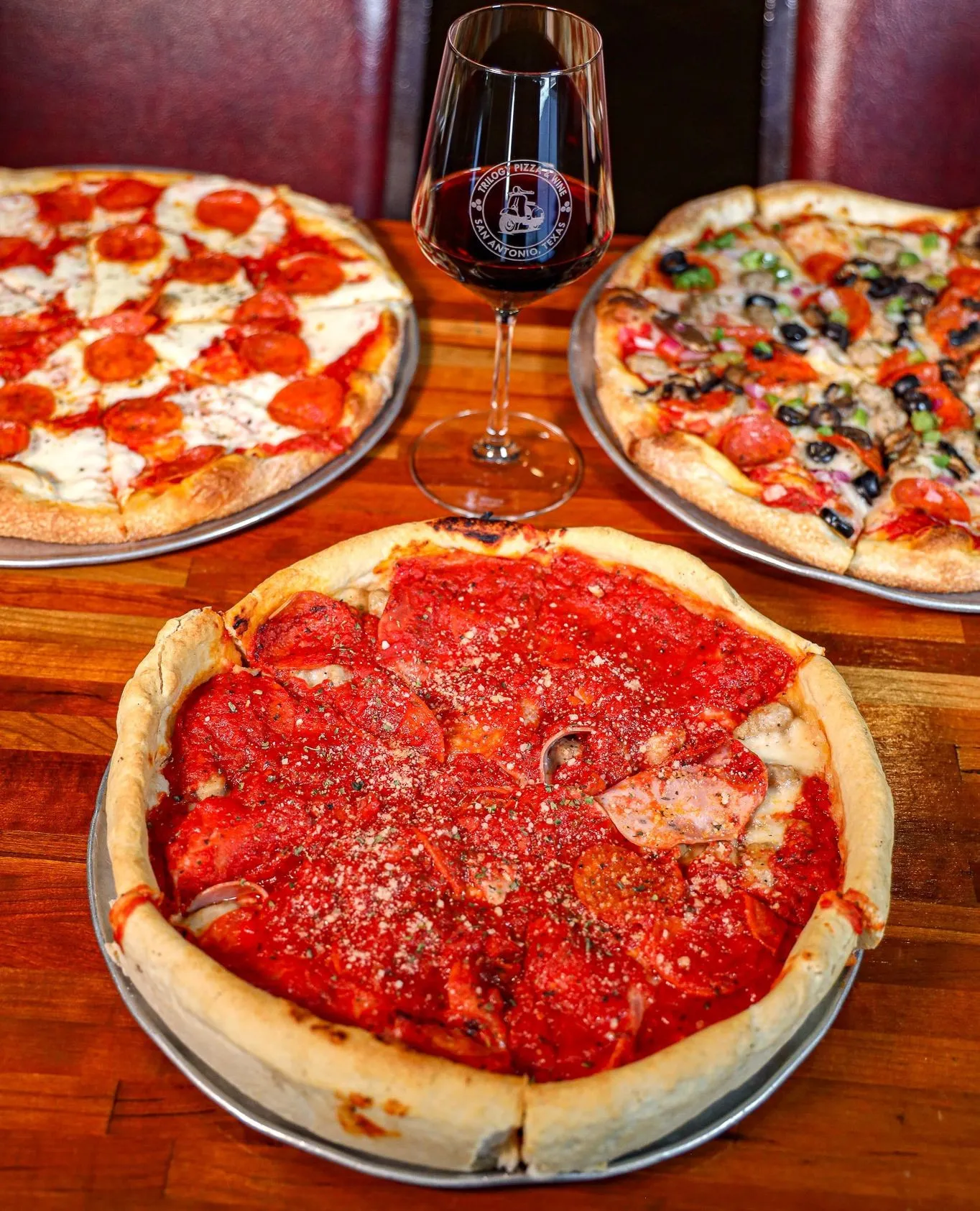 A Chicago deep-dish pizza in front of two thinner pizzas and a glass of wine at Trilogy Pizza