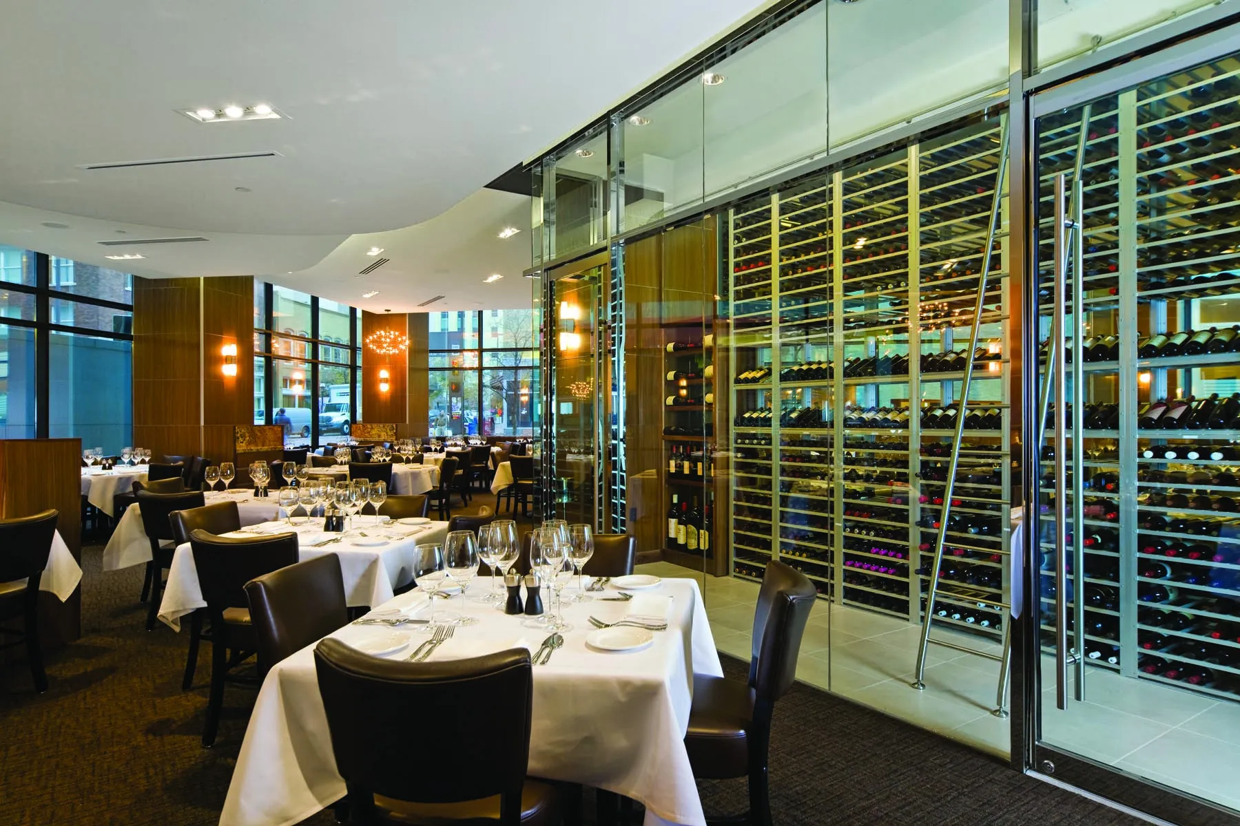 White dining chairs and set tables beside the glass wine wall inside Grace in Fort Worth