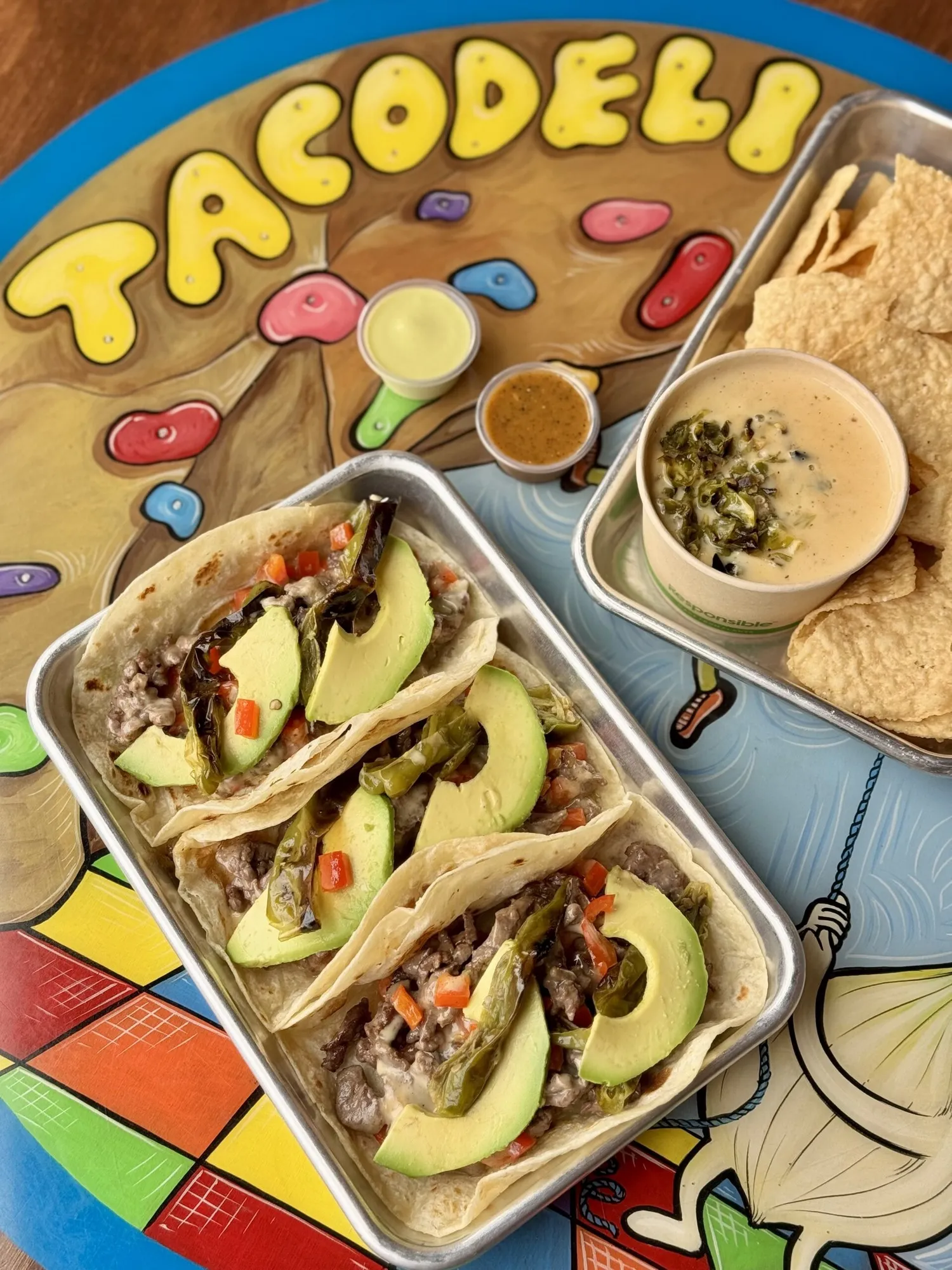 A Tacodeli tray of three tacos with avocado beside queso, chips and salsa on a colorful table