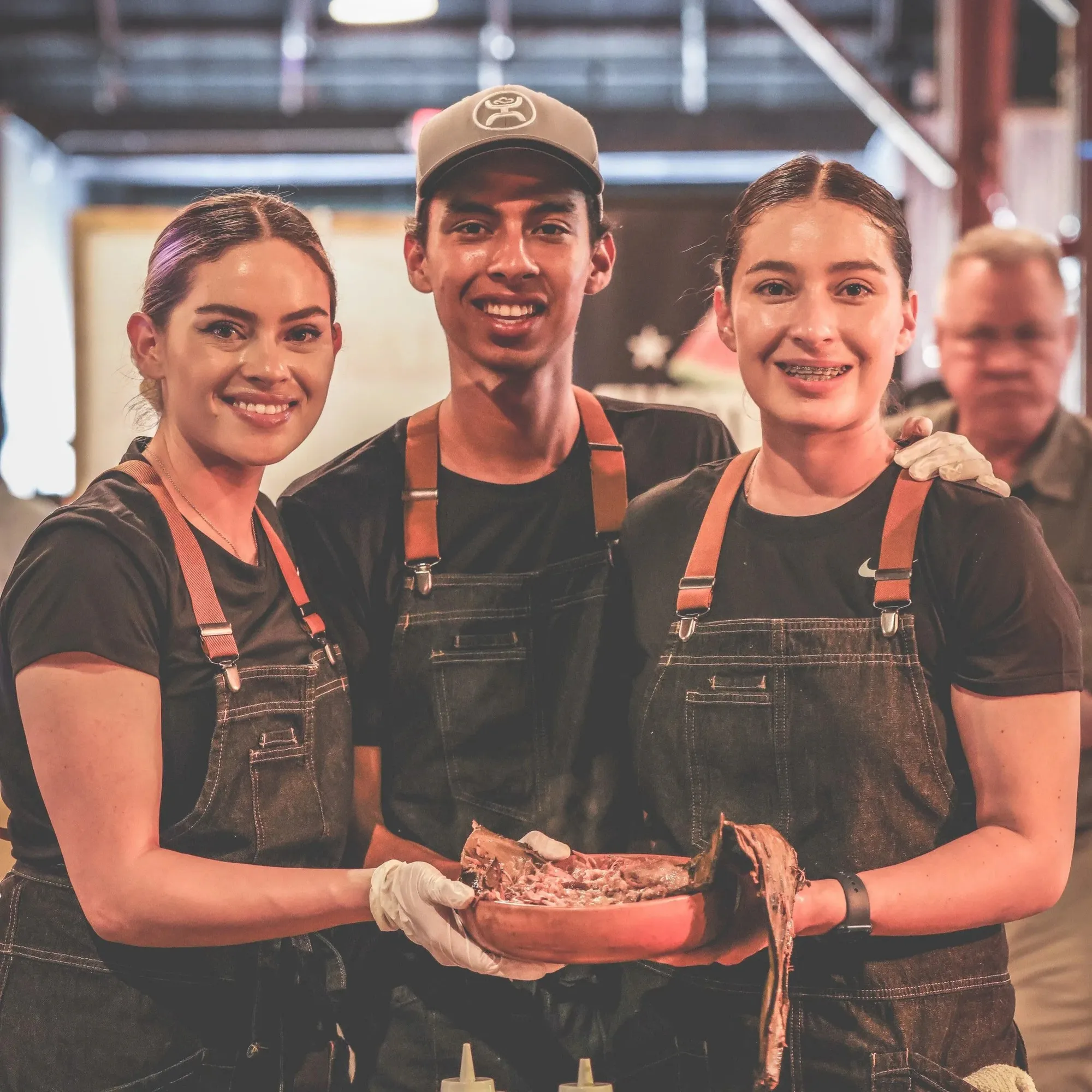 Three members of the La Santa Barbacha team in black aprons, holding a tray of barbacoa