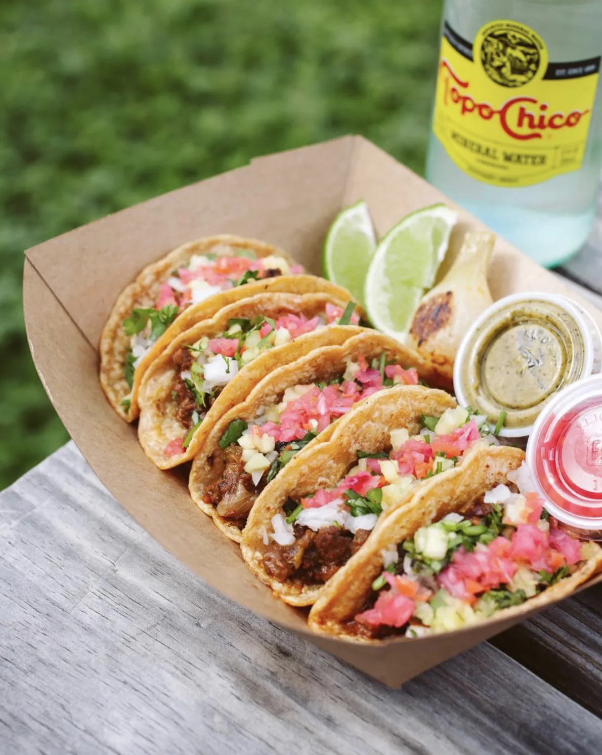 Five small Discada tacos in a cardboard tray with lime, salsa and a bottle of Topo Chico
