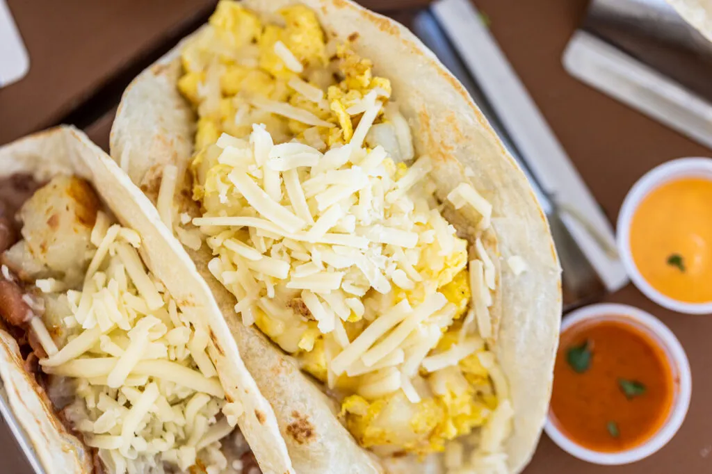 Two Granny's breakfast tacos filled with potato, egg and shredded cheese beside orange and green salsa
