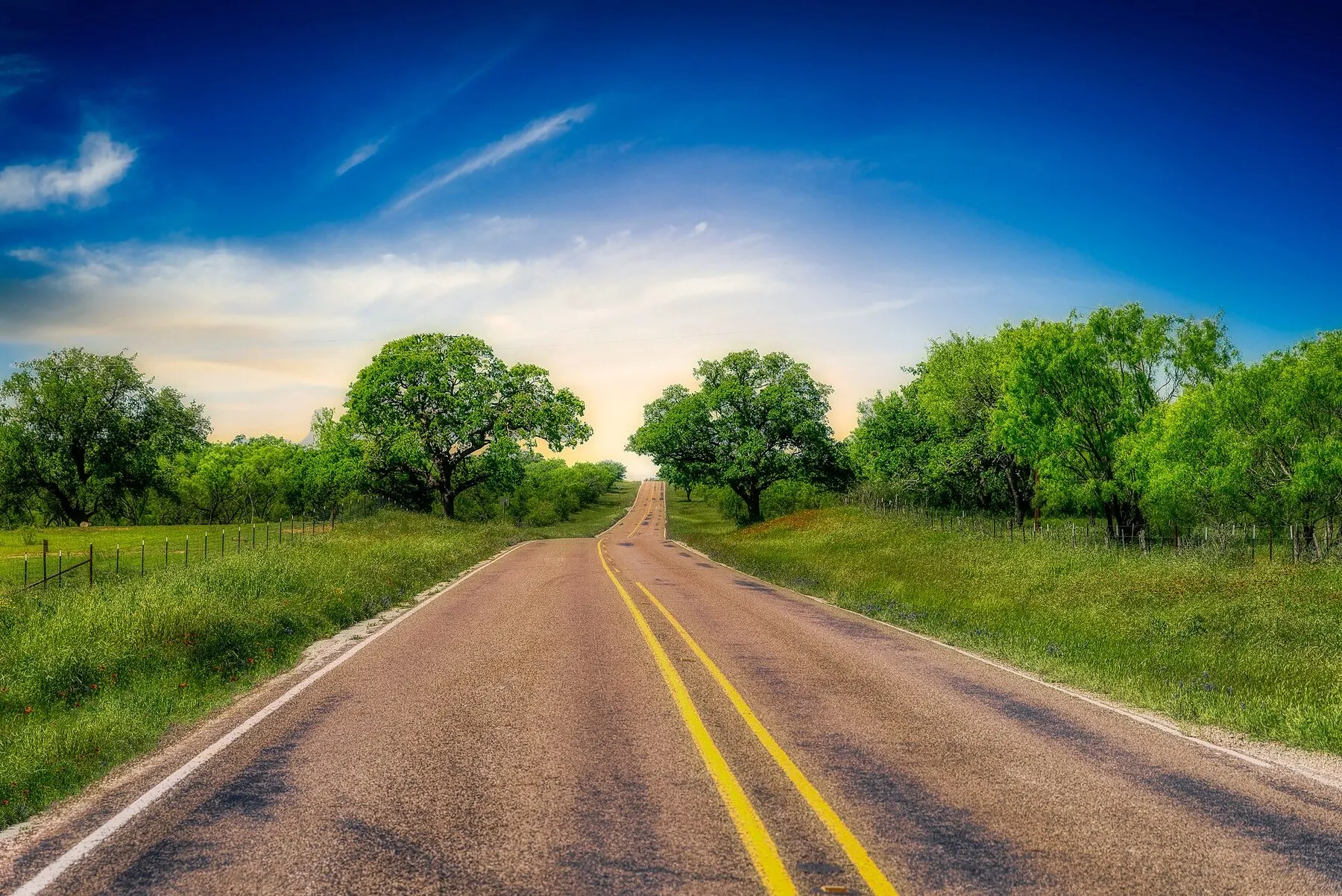 A two-lane road curves through the green Texas Hill Country beneath a broad sky